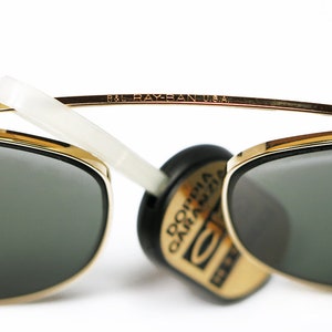 May include: Gold-rimmed sunglasses with dark green lenses. The top bar of the frame is inscribed with "B&L RAY-BAN U.S.A." A white arm extends from the frame. A black and gold tag is attached.