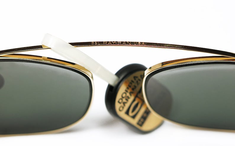 May include: Gold-rimmed sunglasses with dark green lenses. The top bar of the frame is inscribed with "B&L RAY-BAN U.S.A." A white arm extends from the frame. A black and gold tag is attached.