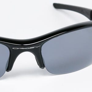 K&ouml;nnte beinhalten: Schwarze Sonnenbrille mit grauen Gl&auml;sern. Die Sonnenbrille hat einen geschwungenen Rahmen und der Markenname "Oakley" ist auf dem Rahmen sichtbar. Auf dem Glas befindet sich ein blau-wei&szlig;er Aufkleber.