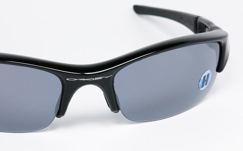 K&ouml;nnte beinhalten: Schwarze Sonnenbrille mit grauen Gl&auml;sern. Die Sonnenbrille hat einen geschwungenen Rahmen und der Markenname "Oakley" ist auf dem Rahmen sichtbar. Auf dem Glas befindet sich ein blau-wei&szlig;er Aufkleber.