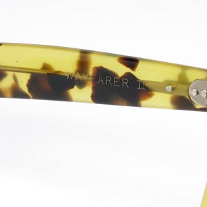May include: Close-up of the arm of Wayfarer II sunglasses. The arm is a translucent yellow-green with dark brown tortoise shell pattern. The text "WAYFARER II" is visible on the arm. The hinge is silver.