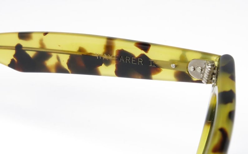May include: Close-up of the arm of Wayfarer II sunglasses. The arm is a translucent yellow-green with dark brown tortoise shell pattern. The text "WAYFARER II" is visible on the arm. The hinge is silver.