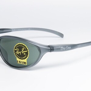 May include: Gray Ray-Ban sunglasses with dark green lenses. The sunglasses have a curved design and the Ray-Ban logo is visible on the lens and the arm. A yellow sticker with text is on the lens.