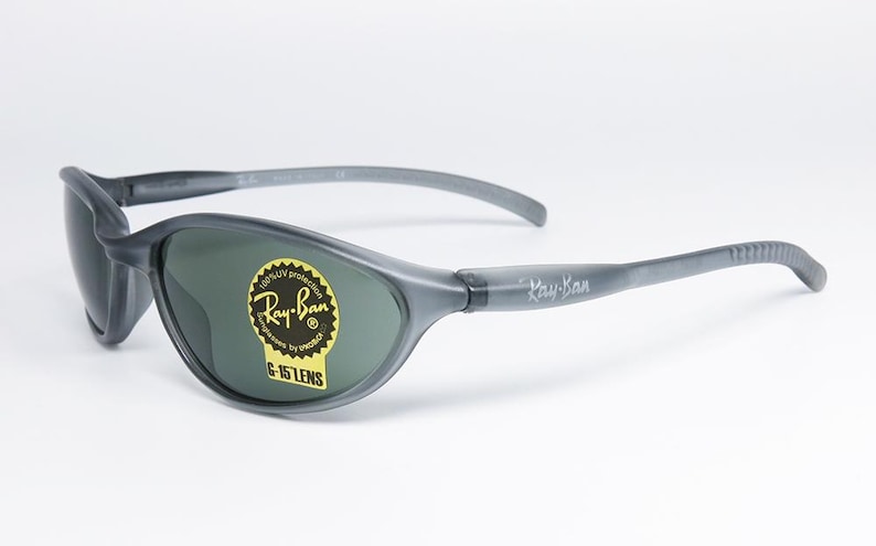 May include: Gray Ray-Ban sunglasses with dark green lenses. The sunglasses have a curved design and the Ray-Ban logo is visible on the lens and the arm. A yellow sticker with text is on the lens.