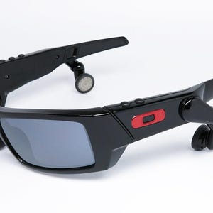 May include: Black sunglasses with gray lenses and integrated audio components. The sunglasses have a sleek, modern design with a red accent on the frame. The earpieces are small and circular.