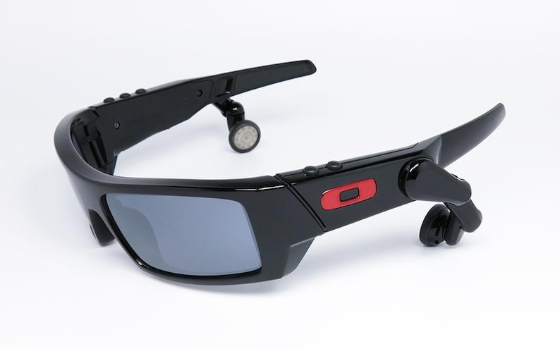 May include: Black sunglasses with gray lenses and integrated audio components. The sunglasses have a sleek, modern design with a red accent on the frame. The earpieces are small and circular.