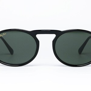 May include: Black sunglasses with oval lenses and a bridge connecting the two lenses. The sunglasses have a thick, glossy black frame and dark green lenses. The Ray-Ban logo is visible on the upper left lens.