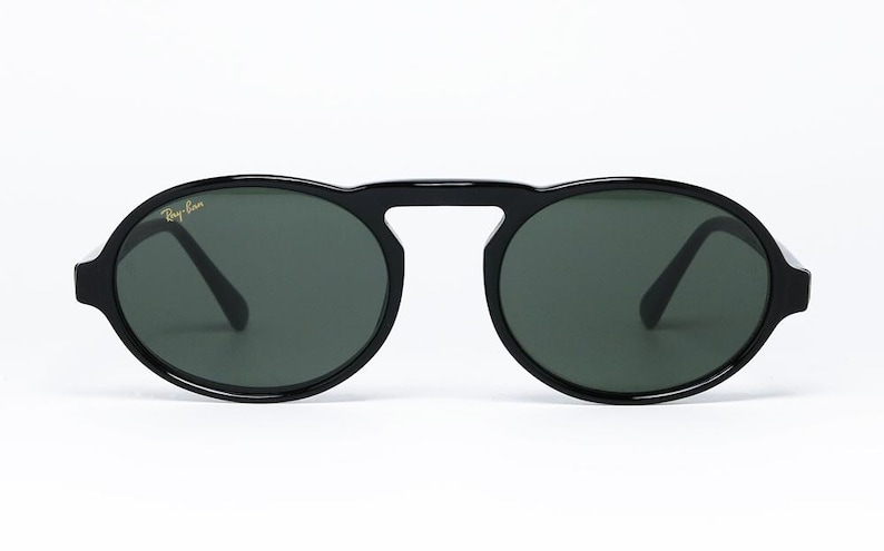May include: Black sunglasses with oval lenses and a bridge connecting the two lenses. The sunglasses have a thick, glossy black frame and dark green lenses. The Ray-Ban logo is visible on the upper left lens.