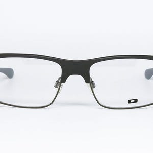 May include: Black rectangular eyeglasses with clear lenses. The frames are a matte black with a subtle, modern design. The temples are a darker gray. The lenses are clear, and the glasses are set against a white background.
