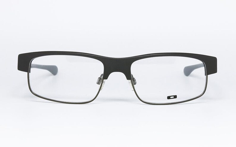 May include: Black rectangular eyeglasses with clear lenses. The frames are a matte black with a subtle, modern design. The temples are a darker gray. The lenses are clear, and the glasses are set against a white background.