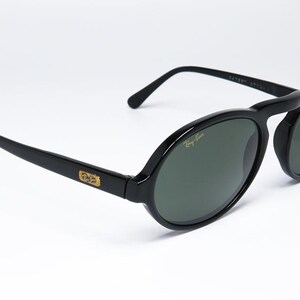 May include: Black oval-framed sunglasses with dark green lenses. The sunglasses have a gold logo on the temples and the Ray-Ban logo on the lens. The frame is glossy and the arms are straight.