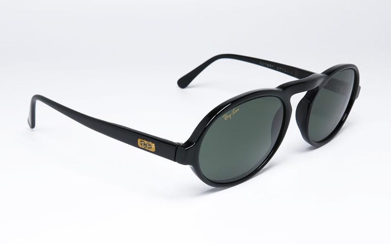 May include: Black oval-framed sunglasses with dark green lenses. The sunglasses have a gold logo on the temples and the Ray-Ban logo on the lens. The frame is glossy and the arms are straight.