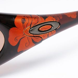 Pode incluir: Close-up de &oacute;culos de sol Oakley com arma&ccedil;&atilde;o castanha e preta. A arma&ccedil;&atilde;o apresenta um vibrante design floral laranja. As lentes s&atilde;o de cor castanho claro e o log&oacute;tipo Oakley &eacute; vis&iacute;vel na haste.