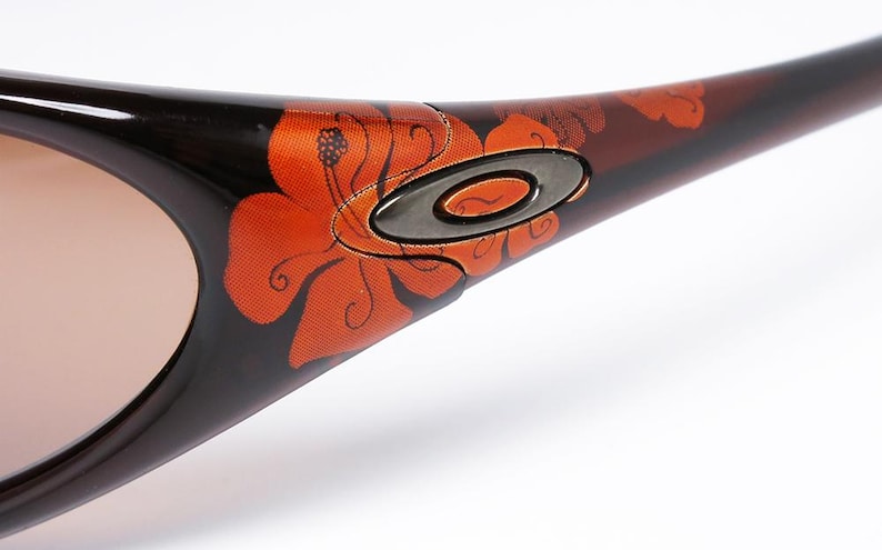 Pode incluir: Close-up de &oacute;culos de sol Oakley com arma&ccedil;&atilde;o castanha e preta. A arma&ccedil;&atilde;o apresenta um vibrante design floral laranja. As lentes s&atilde;o de cor castanho claro e o log&oacute;tipo Oakley &eacute; vis&iacute;vel na haste.