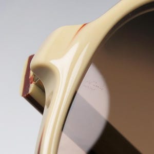 May include: Close-up of sunglasses with a cream-colored frame and dark lenses. The frame has a curved design with a rectangular, maroon-colored accent on the side. The lenses reflect light, creating a gradient effect.