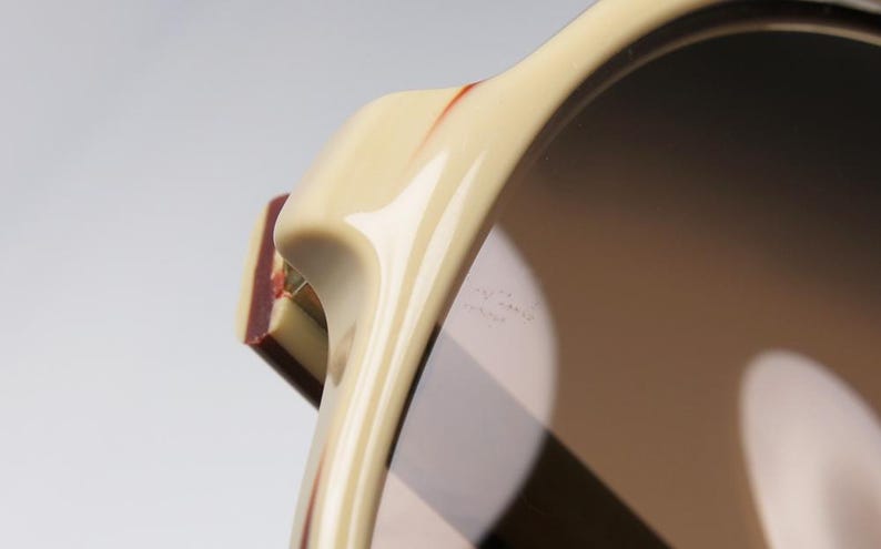May include: Close-up of sunglasses with a cream-colored frame and dark lenses. The frame has a curved design with a rectangular, maroon-colored accent on the side. The lenses reflect light, creating a gradient effect.