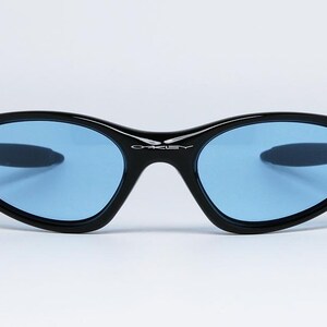 May include: Black sunglasses with blue lenses. The sunglasses have a sleek, curved design with a black frame and the brand name "Oakley" in white on the bridge. The lenses are a light blue color.