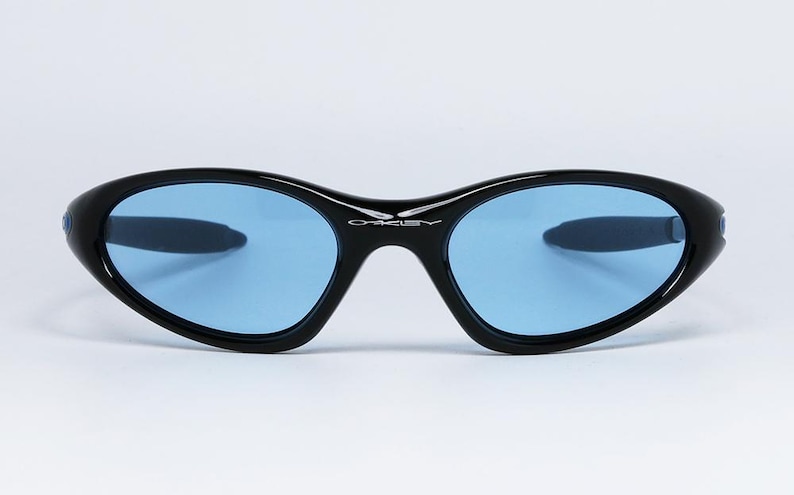 May include: Black sunglasses with blue lenses. The sunglasses have a sleek, curved design with a black frame and the brand name "Oakley" in white on the bridge. The lenses are a light blue color.