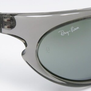 May include: Close-up of gray sunglasses with the Ray-Ban logo in white. The sunglasses have a curved design and a gray tint. The frame is a translucent gray color.