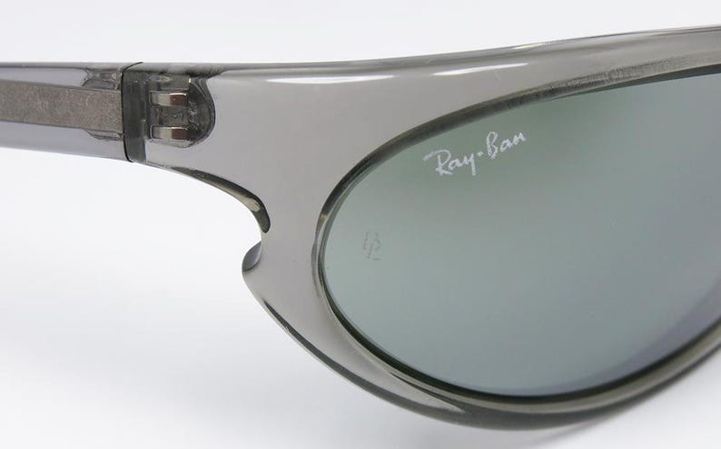 May include: Close-up of gray sunglasses with the Ray-Ban logo in white. The sunglasses have a curved design and a gray tint. The frame is a translucent gray color.