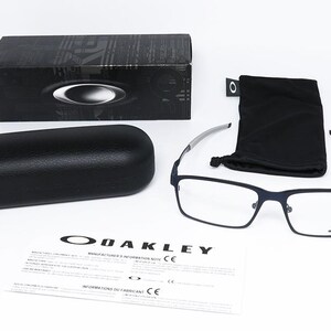 K&ouml;nnte beinhalten: Eine Auswahl an Oakley-Brillenzubeh&ouml;r, darunter eine rechteckige Brille mit marineblauem Rahmen, ein schwarzes Brillenetui, ein schwarzes Mikrofasertuch und eine schwarze Box. Das Oakley-Logo ist auf der Brille, der Box und den Papieren zu sehen.