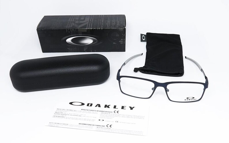 K&ouml;nnte beinhalten: Eine Auswahl an Oakley-Brillenzubeh&ouml;r, darunter eine rechteckige Brille mit marineblauem Rahmen, ein schwarzes Brillenetui, ein schwarzes Mikrofasertuch und eine schwarze Box. Das Oakley-Logo ist auf der Brille, der Box und den Papieren zu sehen.