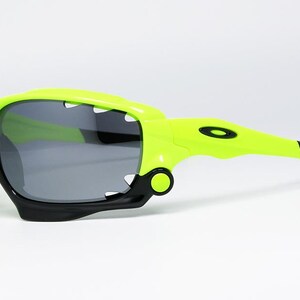 May include: Neon green and black sports sunglasses with gray lenses. The sunglasses have a unique design with a shark-tooth-like frame and a black rubber grip on the temples. The Oakley logo is visible on the temple.