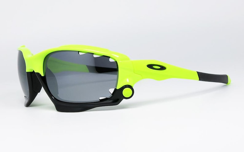 May include: Neon green and black sports sunglasses with gray lenses. The sunglasses have a unique design with a shark-tooth-like frame and a black rubber grip on the temples. The Oakley logo is visible on the temple.