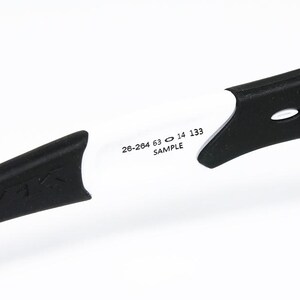 May include: Close-up of a white and black sports eyewear frame. The frame has black temples with a textured design and white arms. The text "26-264 63 14 133 SAMPLE" is printed on the white arm.