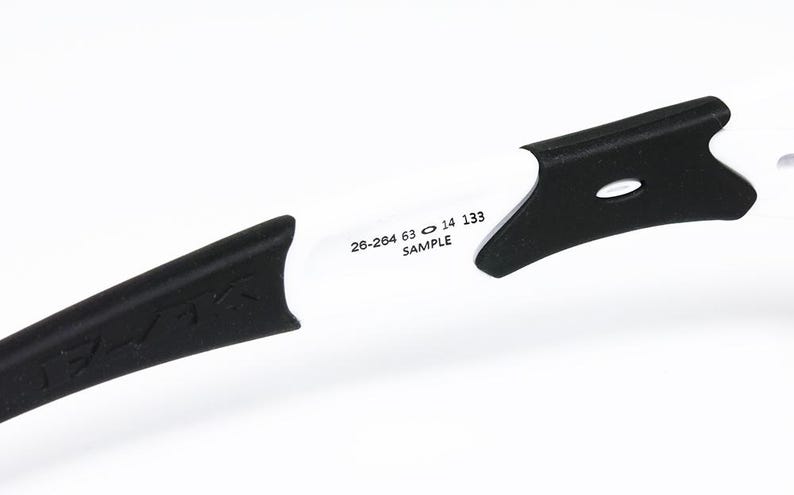 May include: Close-up of a white and black sports eyewear frame. The frame has black temples with a textured design and white arms. The text "26-264 63 14 133 SAMPLE" is printed on the white arm.