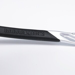 May include: Close-up of a silver and black Radarlock sunglasses frame with the text "RADARLOCK" embossed on the black rubber arm.