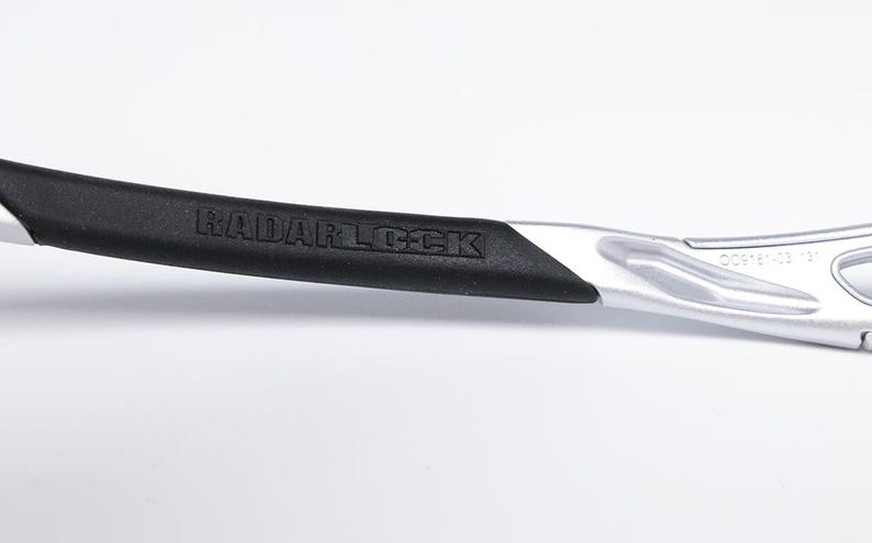 May include: Close-up of a silver and black Radarlock sunglasses frame with the text "RADARLOCK" embossed on the black rubber arm.