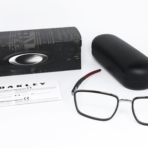 May include: An eyewear set featuring a pair of square-framed glasses with black rims and red accents on the temples. Includes a black case, a black box with the Oakley logo, and a product information sheet. The glasses are designed for vision correction.