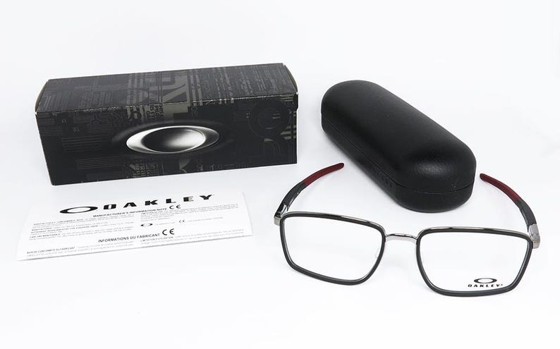 May include: An eyewear set featuring a pair of square-framed glasses with black rims and red accents on the temples. Includes a black case, a black box with the Oakley logo, and a product information sheet. The glasses are designed for vision correction.