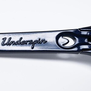 May include: Close-up of the arm of dark blue sunglasses with the word "Underspin" in cursive script. The sunglasses have a metallic finish and a hinge detail. The lenses are a gradient color.