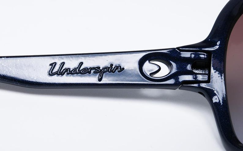 May include: Close-up of the arm of dark blue sunglasses with the word "Underspin" in cursive script. The sunglasses have a metallic finish and a hinge detail. The lenses are a gradient color.