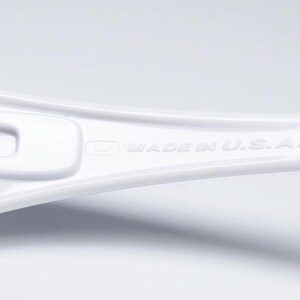 Op de afbeelding: Close-up van een witte brilpoot. De tekst "MADE IN U.S.A." is in reli&euml;f op de poot aangebracht. Het montuur heeft een strak, modern design met een glanzende afwerking. Het CE-keurmerk is ook zichtbaar.