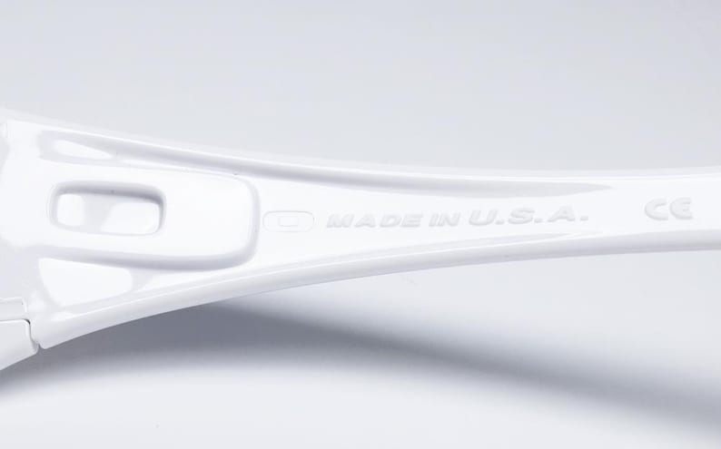 Op de afbeelding: Close-up van een witte brilpoot. De tekst "MADE IN U.S.A." is in reli&euml;f op de poot aangebracht. Het montuur heeft een strak, modern design met een glanzende afwerking. Het CE-keurmerk is ook zichtbaar.