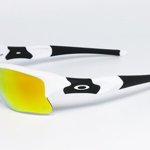 May include: White and black sports sunglasses with a yellow-orange mirrored lens. The sunglasses have a sleek design with black accents and a white frame. The lenses are curved and provide full coverage. The sunglasses are designed for sports and outdoor activities.