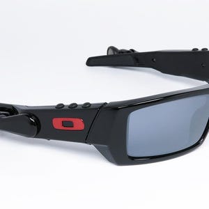 May include: Black sunglasses with gray lenses. The sunglasses have a rectangular frame with a red square logo on the side. The arms have a unique design with a hinge and a curved shape.