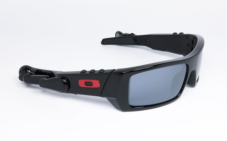 May include: Black sunglasses with gray lenses. The sunglasses have a rectangular frame with a red square logo on the side. The arms have a unique design with a hinge and a curved shape.