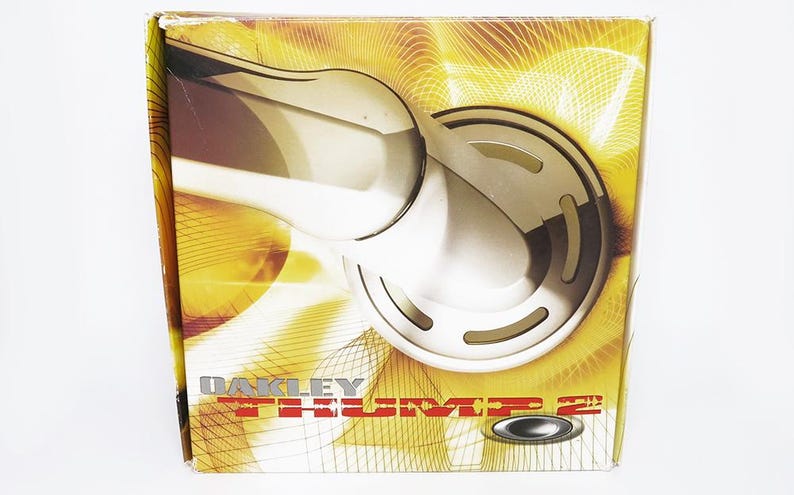 May include: A box with a gold and brown design featuring a silver object. The box has the words "OAKLEY THUMP 2" printed on it. The background has a gold and brown abstract design.