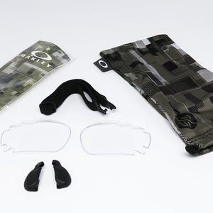 May include: An assortment of Oakley eyewear accessories, including clear lenses, nose pads, a black strap, and a camouflage patterned case and pouch. The Oakley logo is visible on the case and pouch.