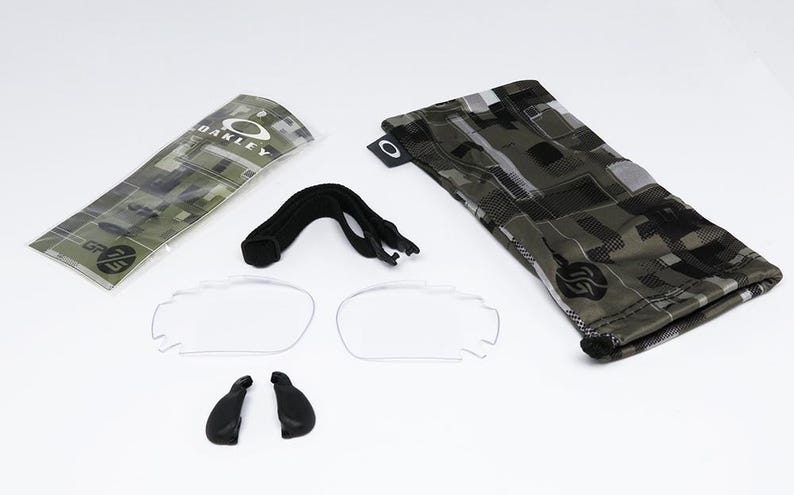 May include: An assortment of Oakley eyewear accessories, including clear lenses, nose pads, a black strap, and a camouflage patterned case and pouch. The Oakley logo is visible on the case and pouch.
