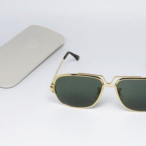 Puede incluir: Gafas de sol con montura dorada y lentes verde oscuro, con un estuche gris claro a juego. Las gafas tienen forma cuadrada y puntas de patillas negras. El estuche es rectangular.
