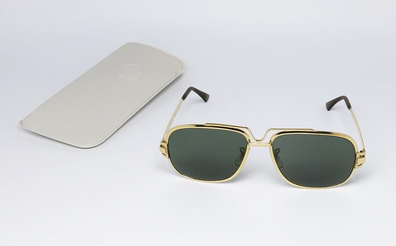 Puede incluir: Gafas de sol con montura dorada y lentes verde oscuro, con un estuche gris claro a juego. Las gafas tienen forma cuadrada y puntas de patillas negras. El estuche es rectangular.