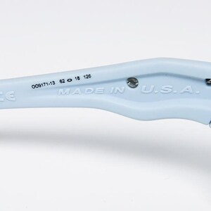 May include: Close-up of a light blue eyeglass arm with a black tip. The text "MADE IN U.S.A." is printed on the arm, along with other numbers and the CE mark. The arm is attached to a gray hinge.