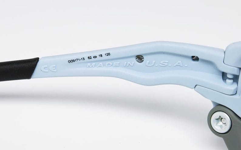 May include: Close-up of a light blue eyeglass arm with a black tip. The text "MADE IN U.S.A." is printed on the arm, along with other numbers and the CE mark. The arm is attached to a gray hinge.