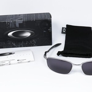 May include: A pair of sunglasses with a silver frame and dark lenses, accompanied by a black carrying pouch and a black box. The box features a logo and text. A white card with text is also present.