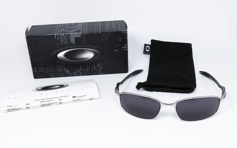 May include: A pair of sunglasses with a silver frame and dark lenses, accompanied by a black carrying pouch and a black box. The box features a logo and text. A white card with text is also present.